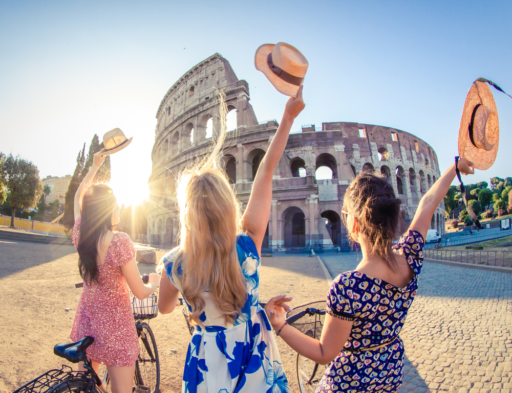 Three girls tip their hats and wear sun dresses as the sun shines over the Colosseum in Italy in May. 