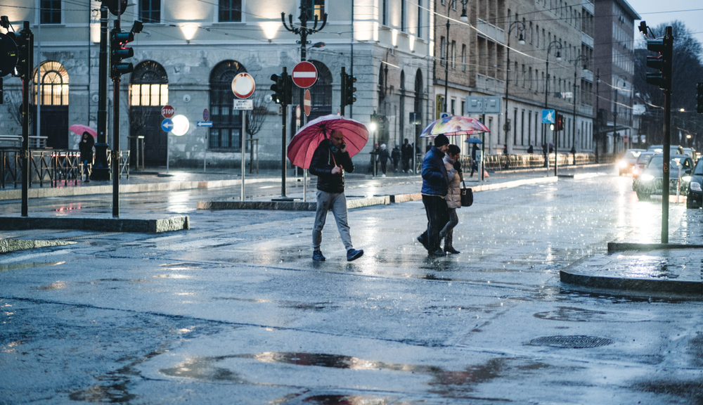 Three people cross the street in Italy in May with umbrellas as the rain pours down and reflects off the road.