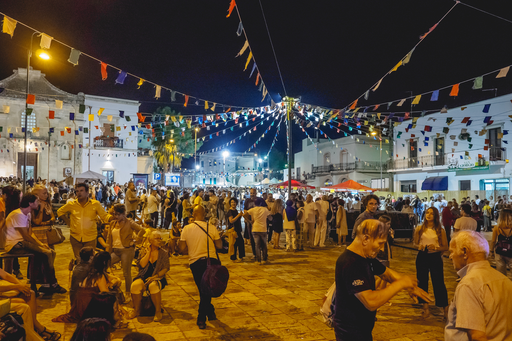 Groups of tourists and locals gather at a festival, eating, dancing and talking under brightly colored flags. This is during the harvest season. 