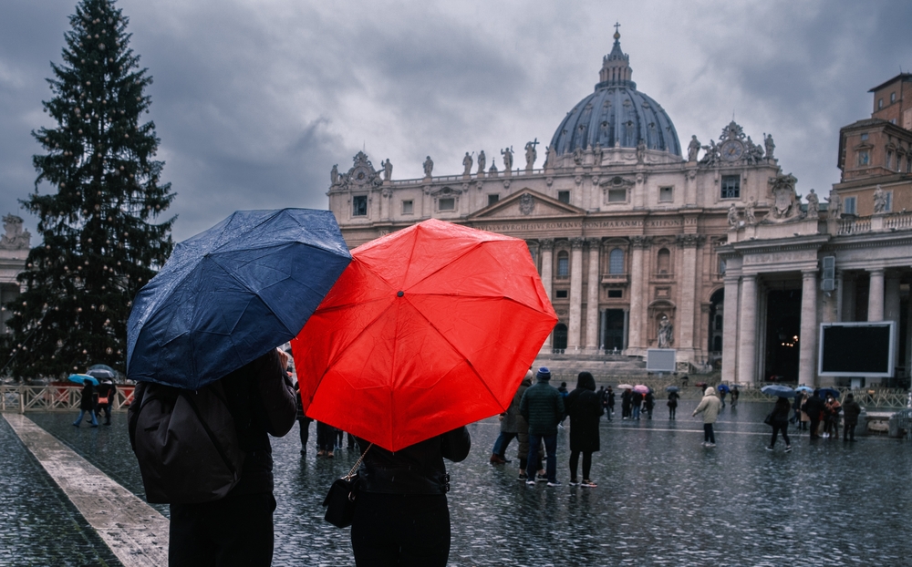 A couple with a red and blue umbrella look at a bolding as it rains: they are dressed in rain jackets and warm layers to combat the autumn rain and cooler weather. 