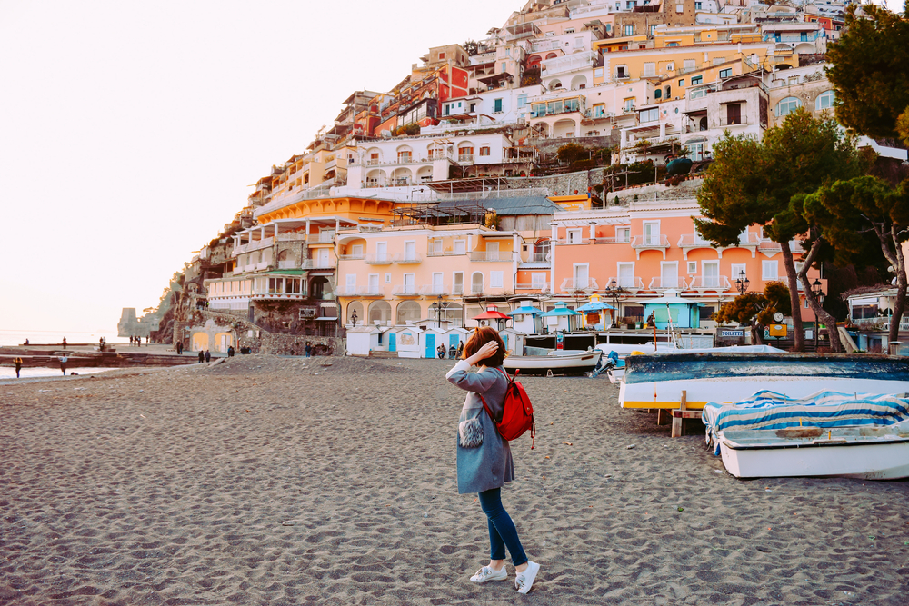 A lone woman in long sleeves carries a backpack as she looks out over the water on the coast of Cinque Terre. It is common for there to not be many people near the water in Italy in October.