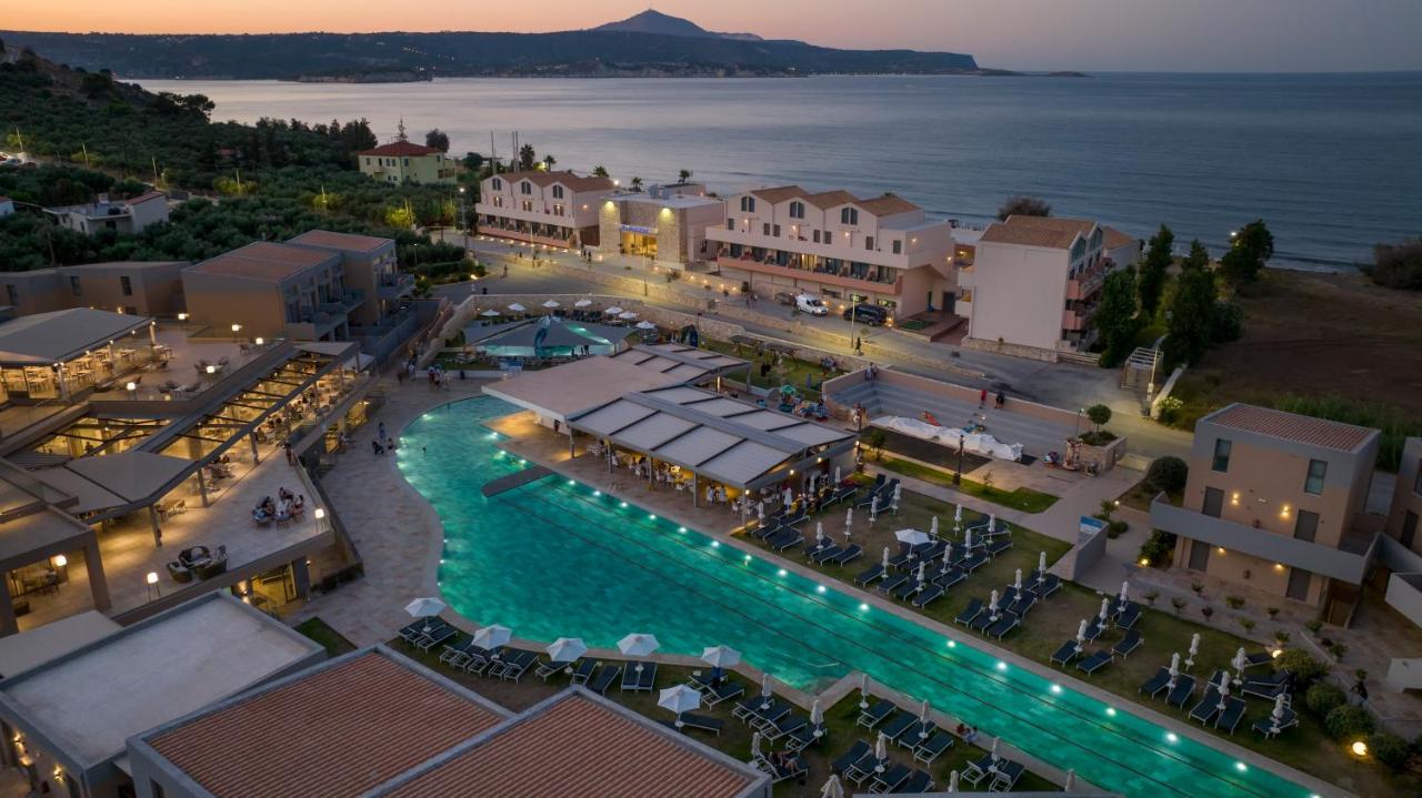 An aerial view of a resort in Greece on the beach with a large pool, villas, and other buildings at sunset