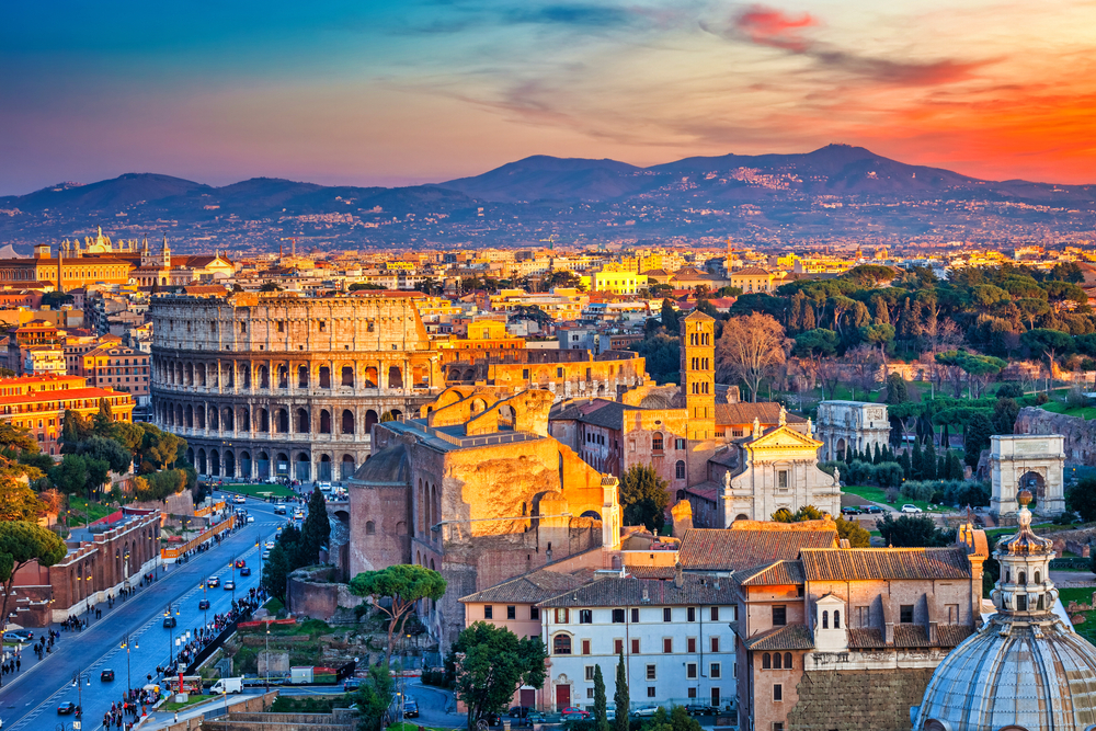 View looking out over Rome at sunset featuring the Colosseum and mountains in the distance.