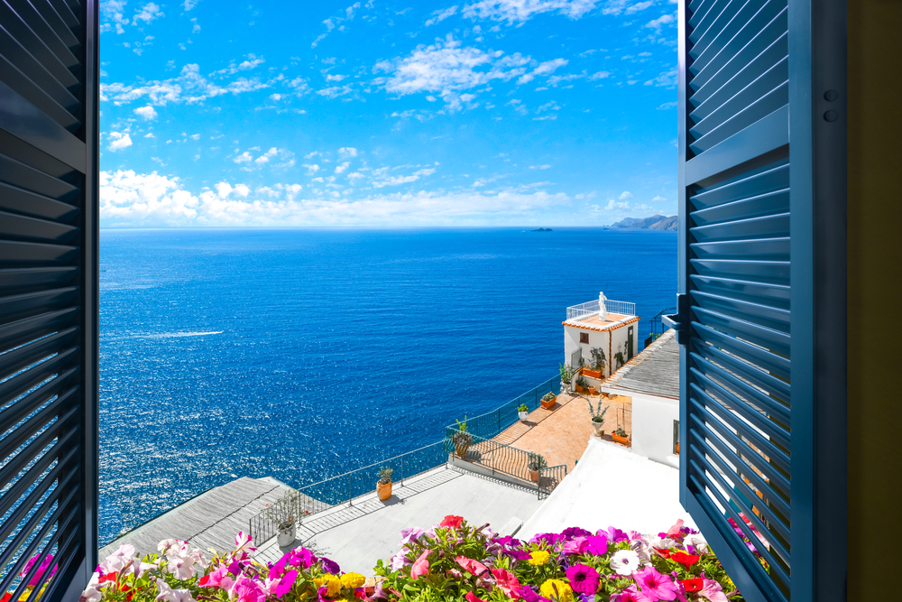 Village of Manarola, on the Cinque Terre coast of Italy with flowers, Ocean viewed out of window with blue shutters.