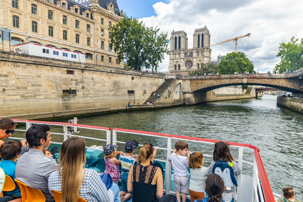 Groups of people gather at the cusp of a boat cruise on the seine river: young children stand close to the railings and peer out at Notre Dame in the distance. 