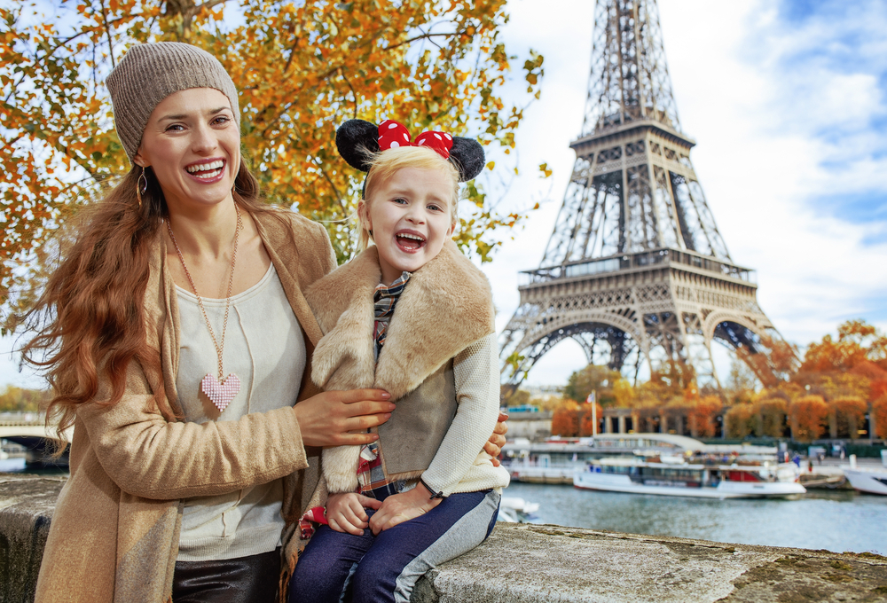 A young girl wears Minnie ears while posing with her mom in front of the Eiffel Tower after a "we're going to Disneyland Paris!" reveal.