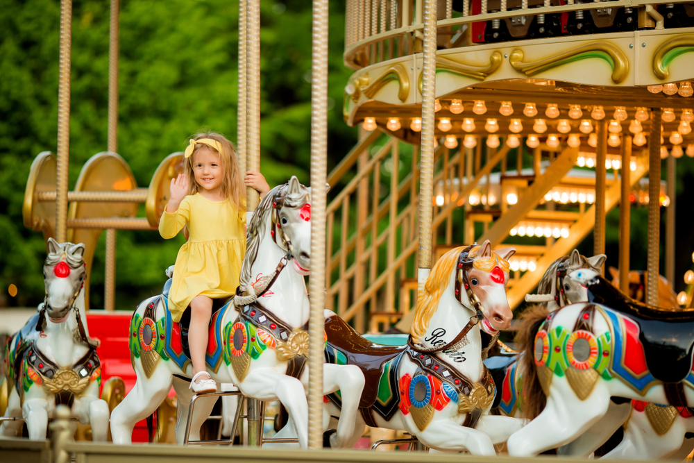 A young girl in a yellow dress waves at someone from her horse on the carousel in the middle of a Paris park.