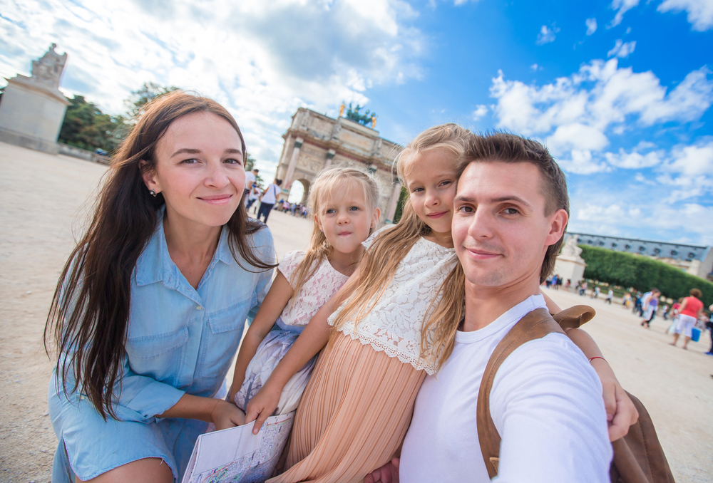 A family of four-- which features two young girls-- stand in front of the Louvre waiting for a tour to start. One of the girls clutches a map.