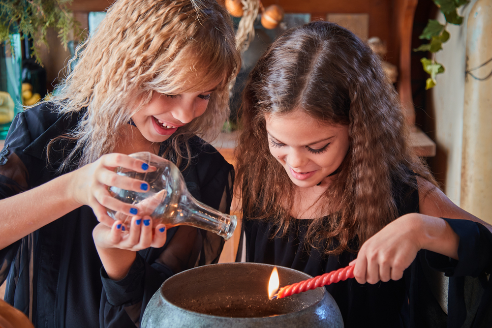 Two young girl sin all black and crimped hair lean over a bowl and pour scenes into it to create a fragrance. 