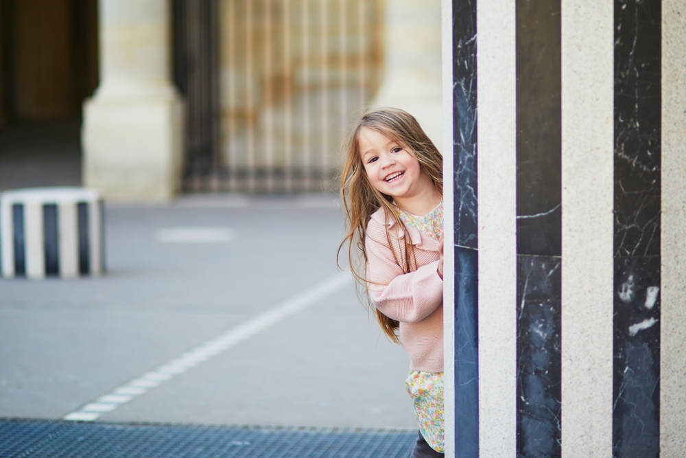 A young girl with a big smile peaks around a column at the Paris Royal Garden. 