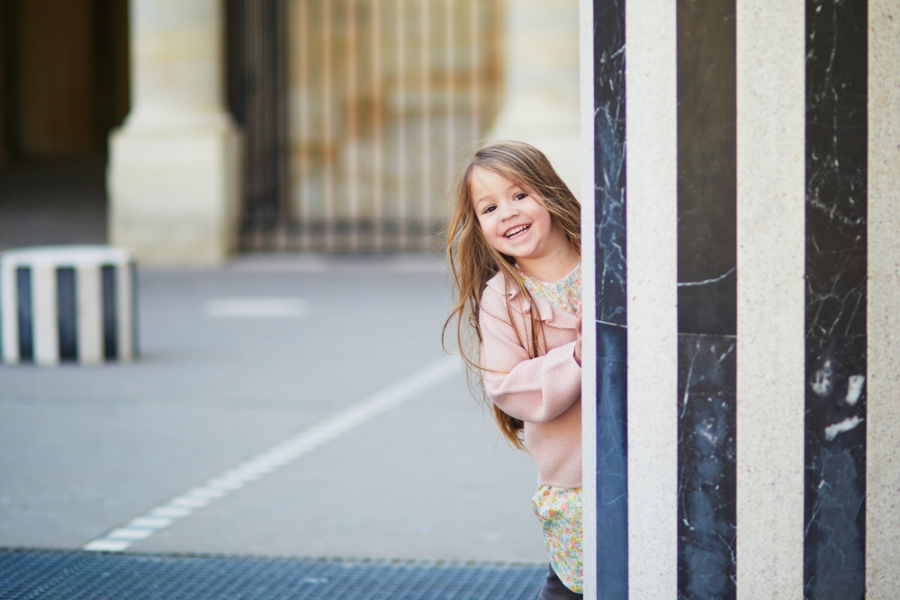 A young girl with a big smile peaks around a column at the Paris Royal Garden.
