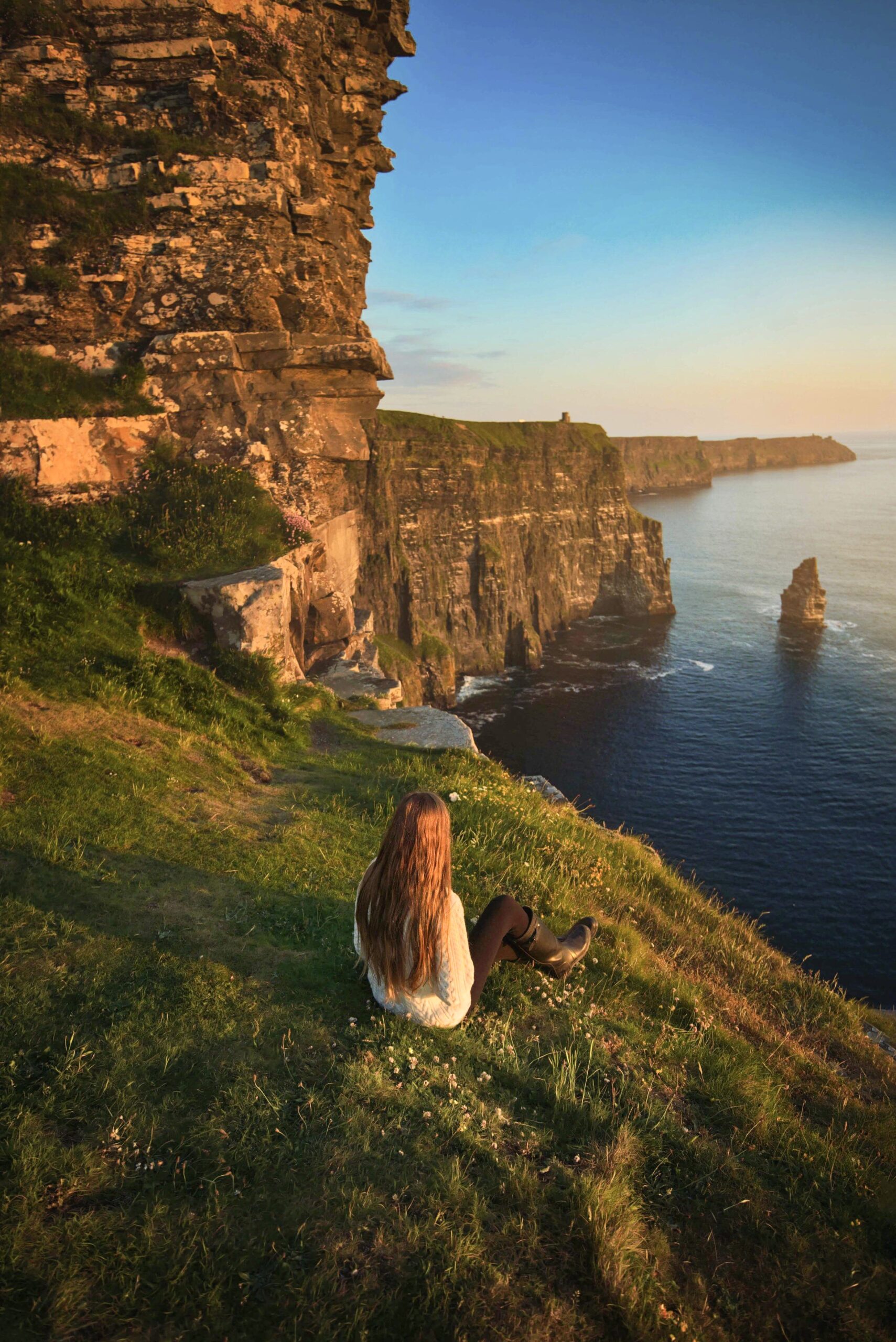 Woman sitting in grass overlooking the epic Cliffs of Moher at sunset.