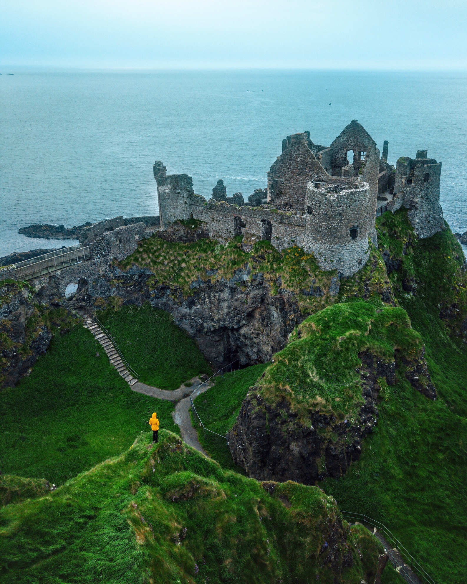 Figure in yellow standing on a cliff looking at Dunluce Castle on a cloudy day.
