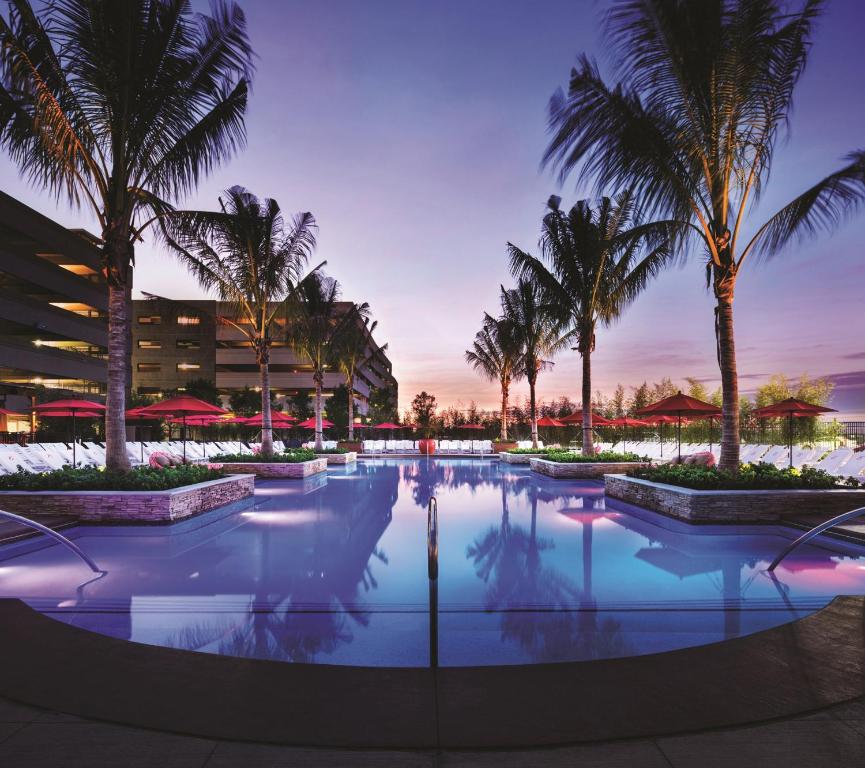 A pool at dusk surrounded by palm trees and red umberellas. The hotel building is in the background. 