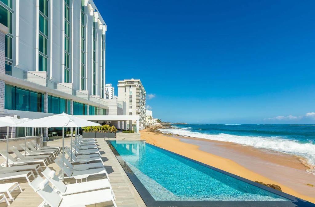 Rectangle pool over the beach in front of a large modern white hotel. 