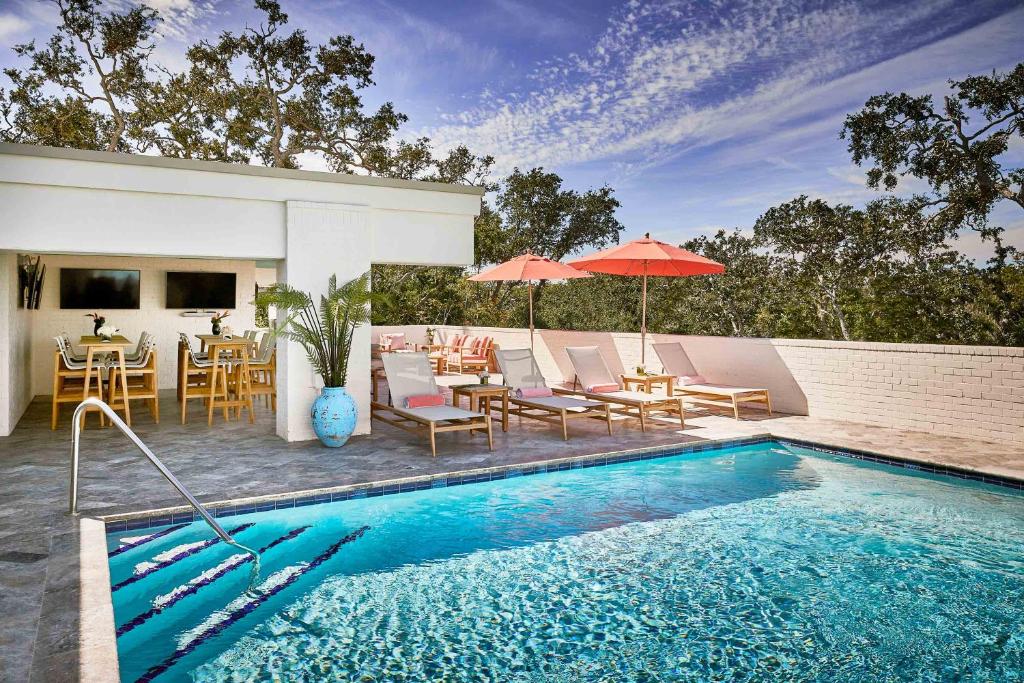 A hotel pool surrounded by loungers and tables and chairs. Trees are in the background. 