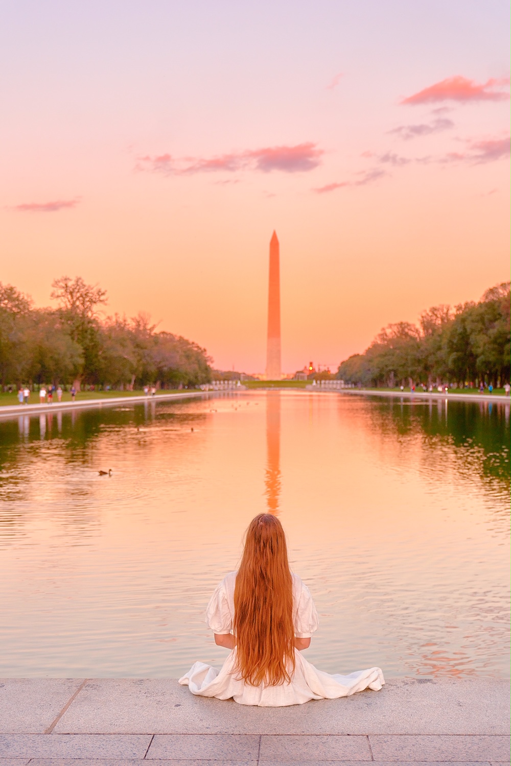 Woman with long hair sitting on  the edge of the reflecting pool in front of the Washington Monument in Washington DC.