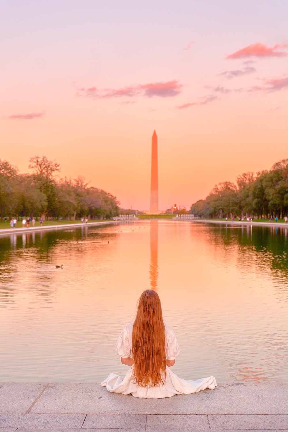 Woman with long hair sitting on the edge of the reflecting pool in front of the Washington Monument in Washington DC.