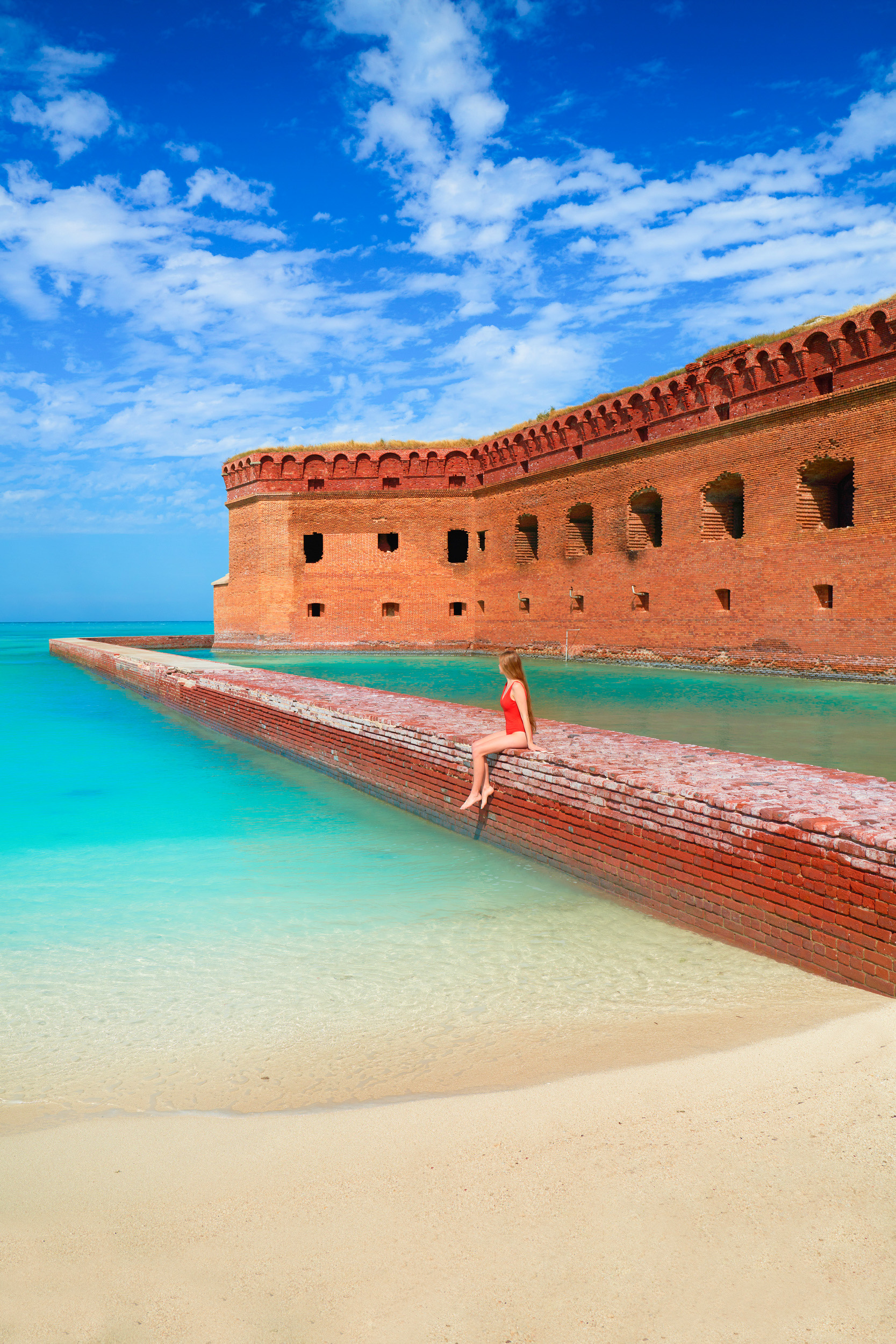 One of the BEST things to do in Florida is a visit to Dry Tortugas Fort Jefferson.  A girl in red swimsuit sits on red brick wall with turquoise waters.