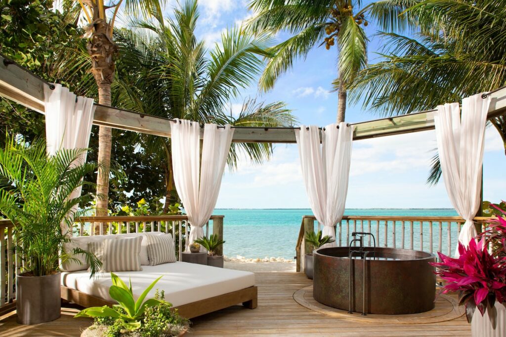 Little Palm Island is one of the all inclusive resorts in Florida, shown here is a wooden patio featuring a daybed, copper tub surrounded dby palm trees with ocean views framed by white curtains from the room