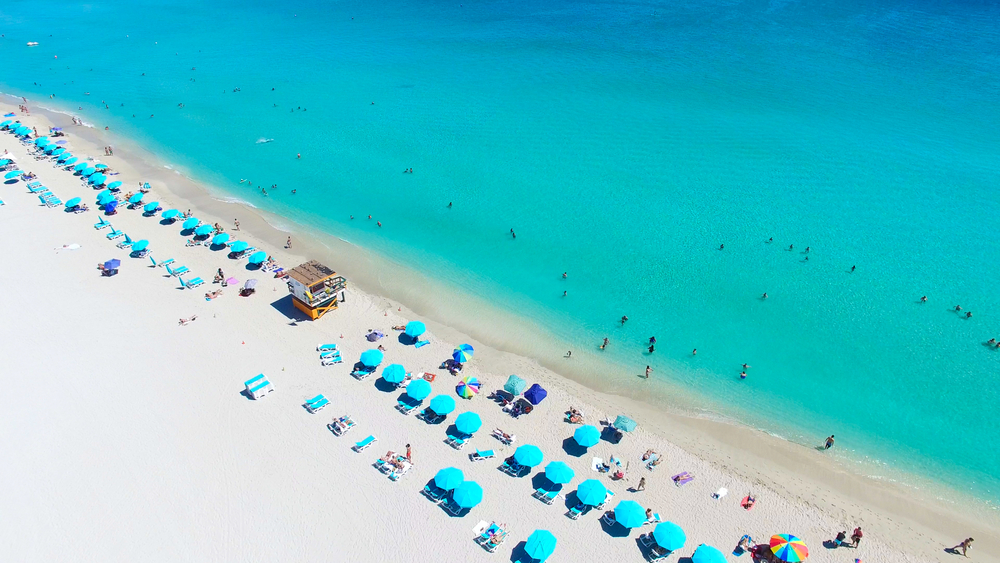 The iconic south beach from above with white sand beach filled with teal beach umbreallas with turquoise waters filled with swimmers