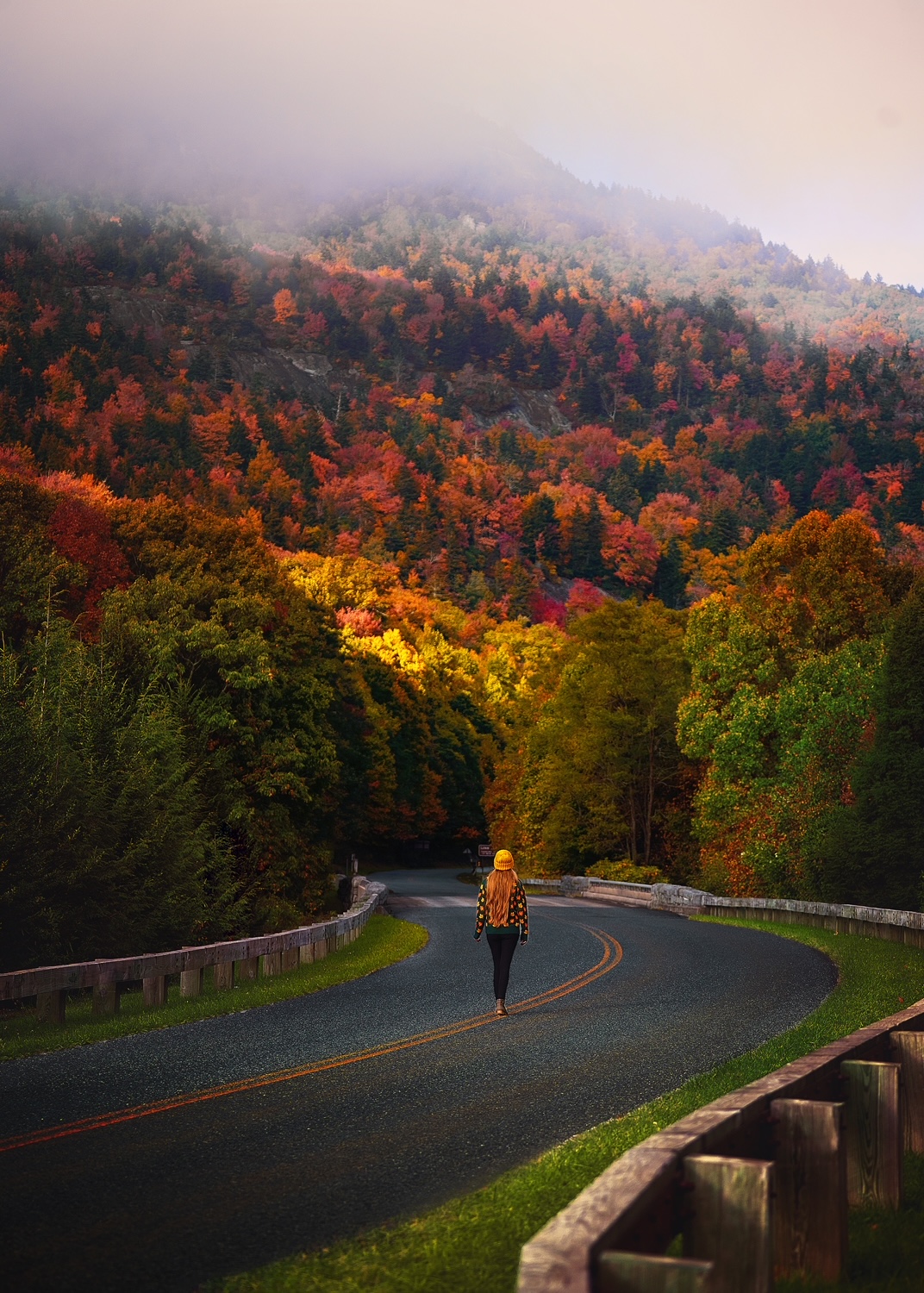 Woman in a sweater walks along a road under a mountain with colorful fall foliage in the USA.