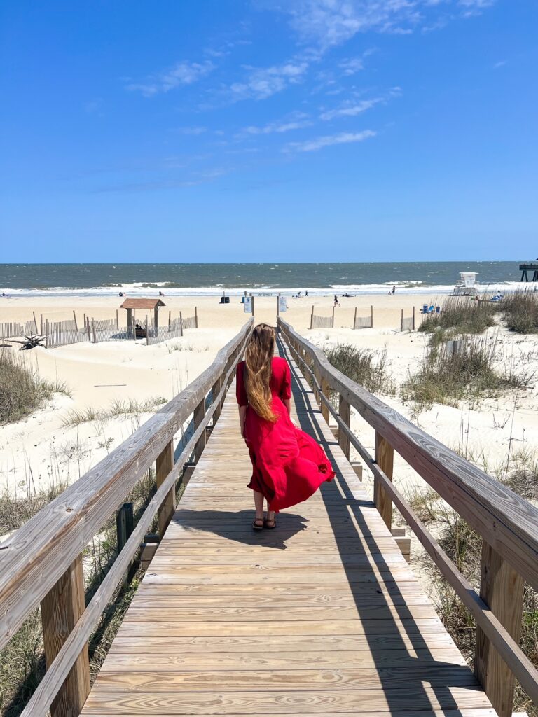 a girl in red dress walking on the wooden boardwalk to the beach