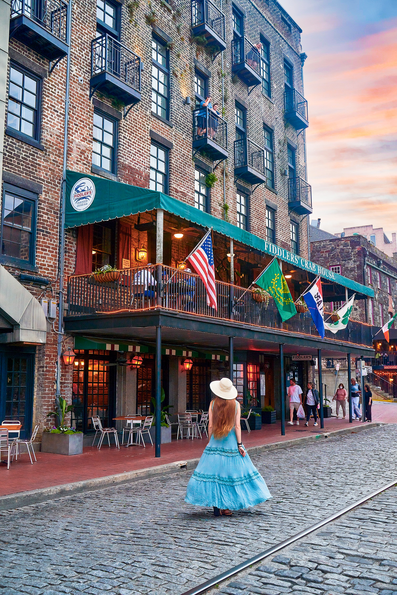 a girl in a blue dress and sunhat walking down the cobblestone street along Riverfront with a two story historic building  in Savannah Ga