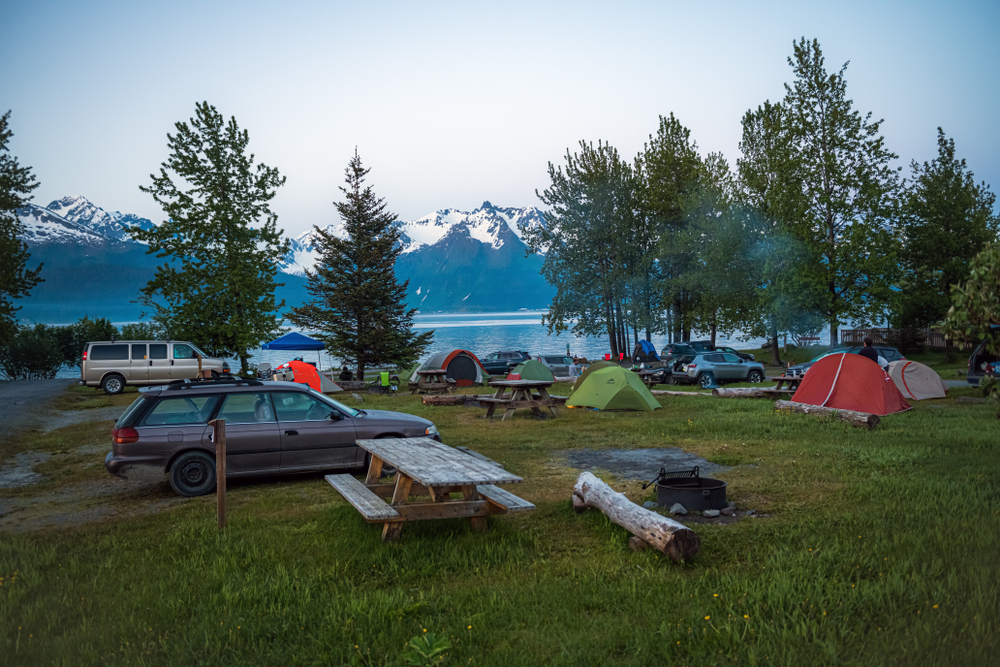 A serious of cars and tents park by the water in Alaska: you can see snow capped mountains in the back, making it a must see location for the best beach camping in the USA. 