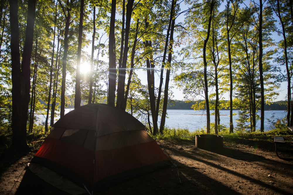 A tent hidden behind trees peers out over the water and rising sun in Michigan. Despite it not being an ocean, the Great Lakes are one of the top areas for the best beach camping in the USA.