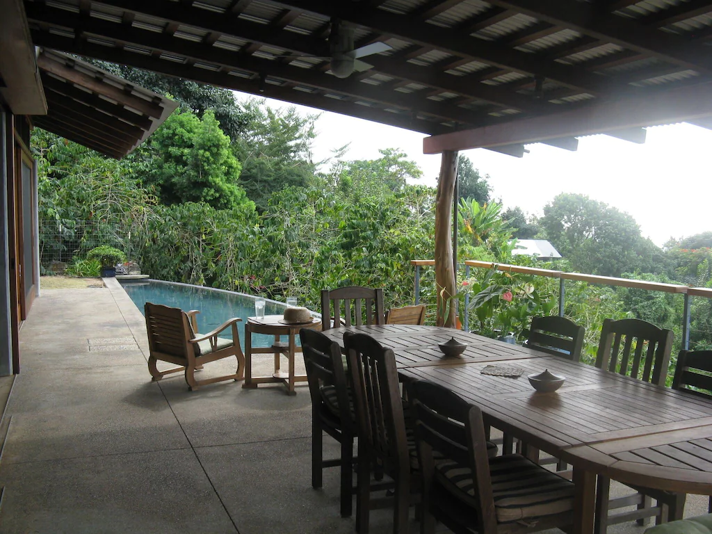 View of the lap pool and large outdoor dining table surrounded by lush greenery at Mo Snell's Coffee Farm House