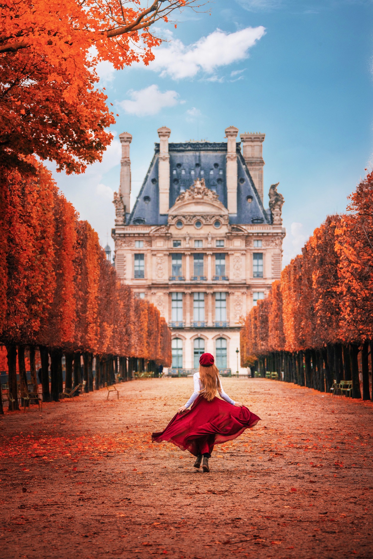 Woman in flowing red shirt and beret walks down a path of orange trees in fall in Paris.