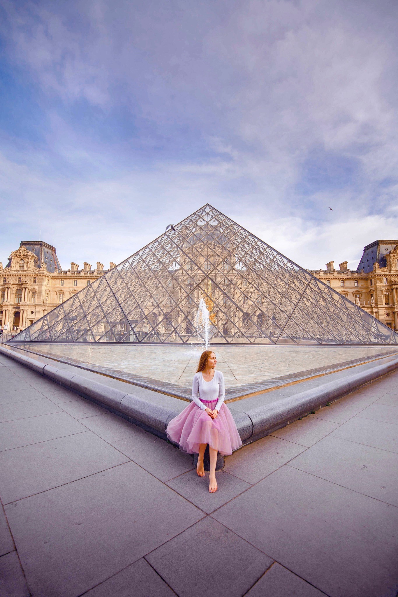 Woman in pink skirt sitting on the edge of the fountain surrounding the iconic glass pyramid at the the Louvre.