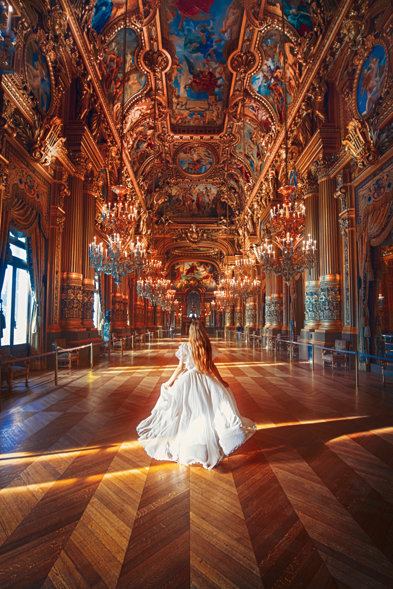 Woman in white dress walking through hall full of chandeliers in Palais Garnier, one of the most beautiful places in Paris.