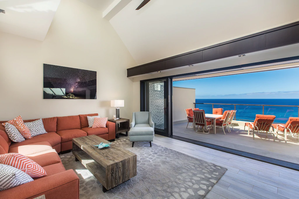 View of a comfortable living room and deck with orange accents and a stunning view of the ocean. 