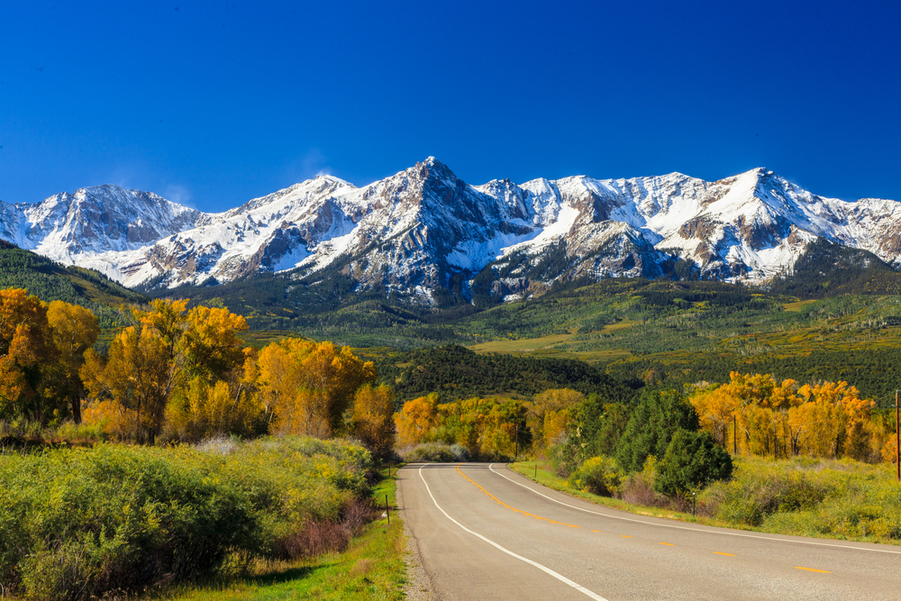 Road heading towards fall trees and snow capped mountains.