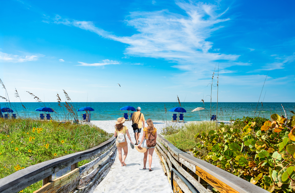 A family of three cross a boardwalk onto the white sand of Treasure Island. The blue skies and teal water stretch in front of them.