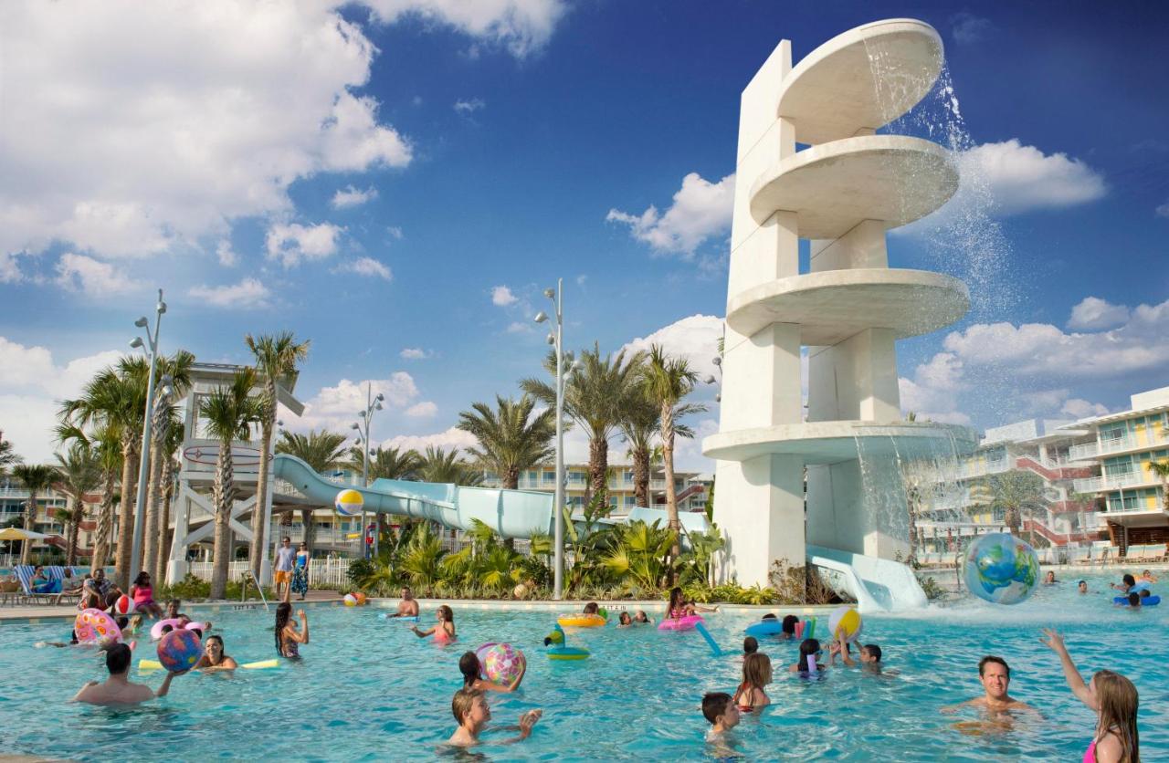A tall tower of waterfall with a water slide and a lagoon pool with children playing with beach balls and swimming in the foreground