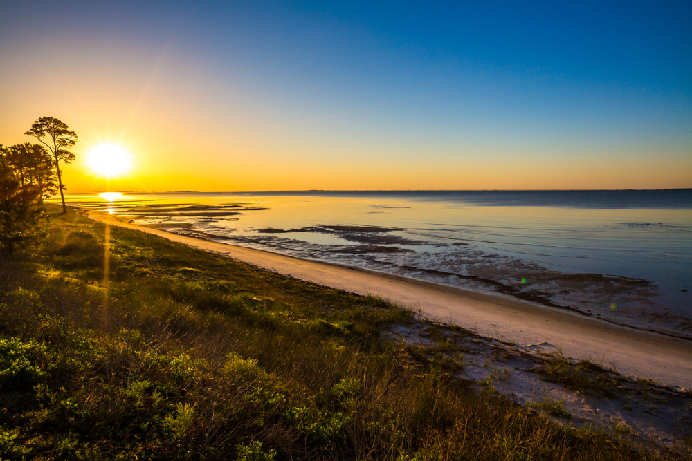 a dusky sky with dark blue skies and bright yellow sun setting into an orange horizon and shining on a clean beach and deep green grass at one of the best small towns in Florida