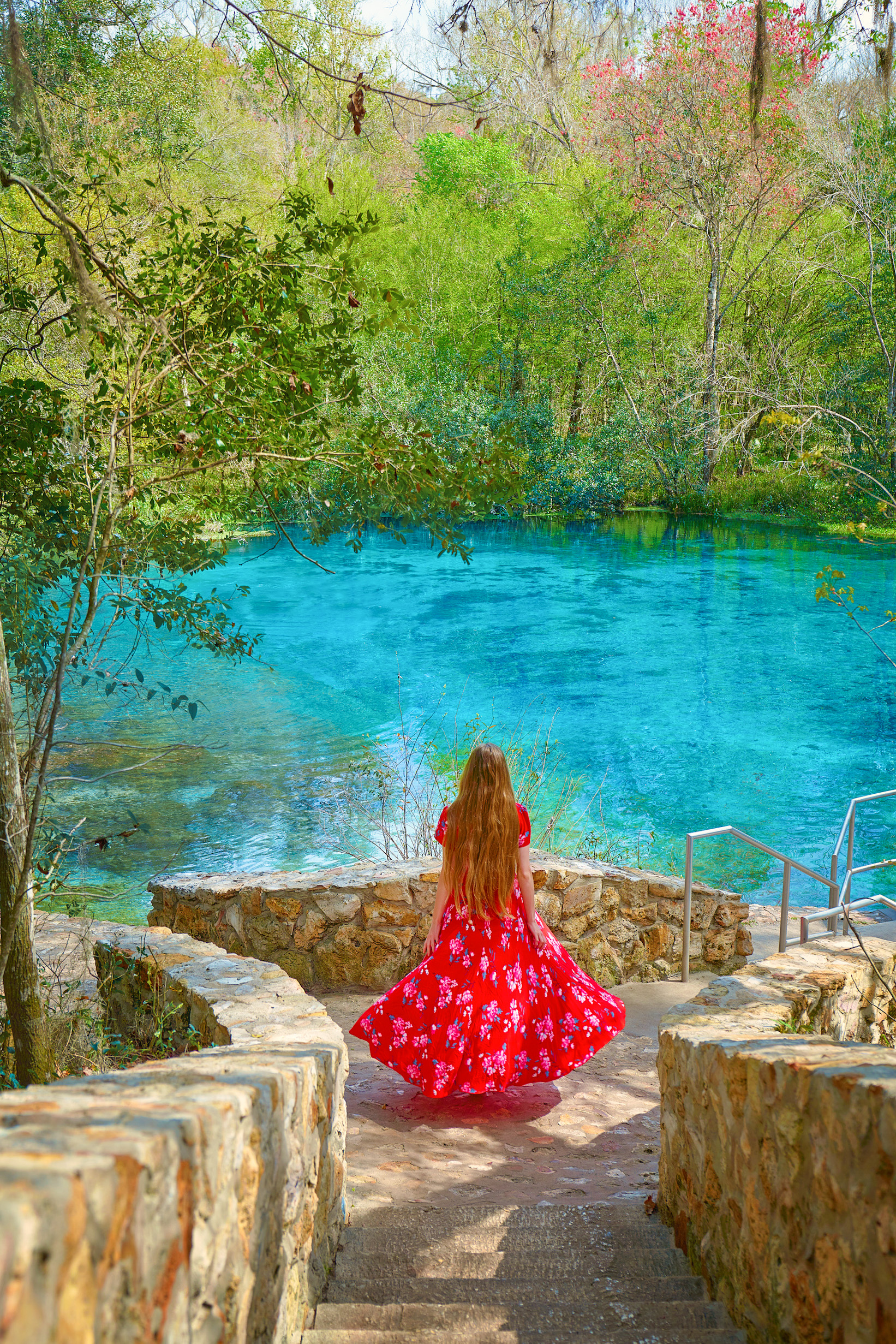 a woman in a bright red dress with floral pattern stands on a rocky pathway leading down to bright blue water of a natural spring at one of the best small towns in Florida!