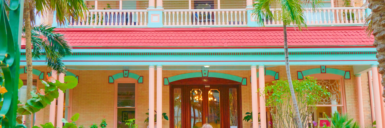 a bright coral pink, yellow and light turquoise coloured house with bright green palm trees and bushes with a woman standing out front of the building in a long floral dress in one of the best small towns in Florida