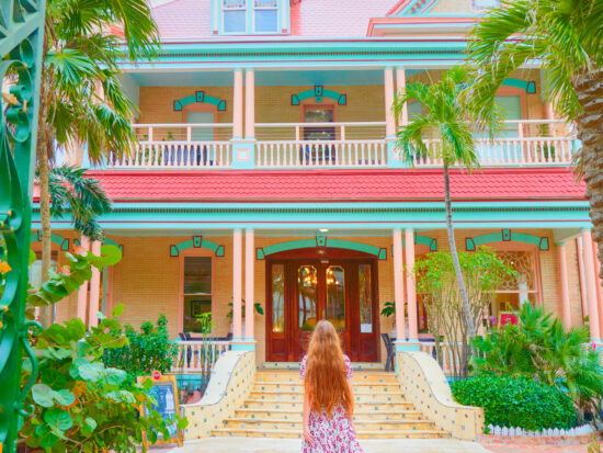 a bright coral pink, yellow and light turquoise coloured house with bright green palm trees and bushes with a woman standing out front of the building in a long floral dress in one of the best small towns in Florida