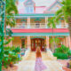 a bright coral pink, yellow and light turquoise coloured house with bright green palm trees and bushes with a woman standing out front of the building in a long floral dress in one of the best small towns in Florida