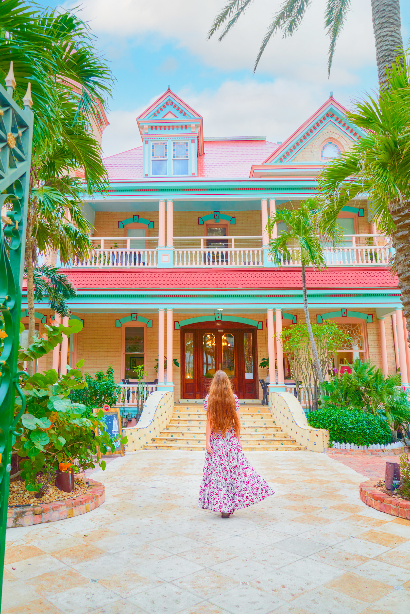 a bright coral pink, yellow and light turquoise coloured house with bright green palm trees and bushes with a woman standing out front of the building in a long floral dress in one of the best small towns in Florida