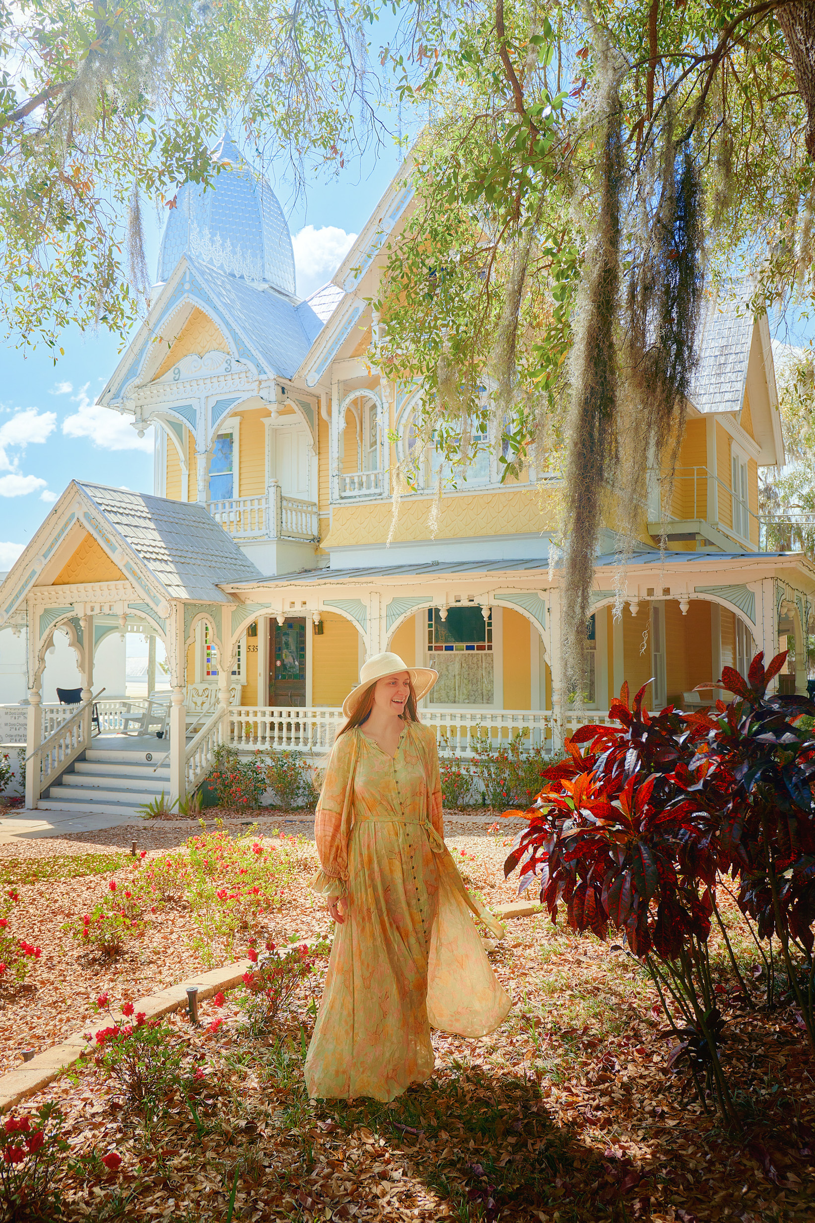 woman in greenish yellow dress with orange accents and wide brimmed hat stands out front of another pastel coloured house with yellow and blue hues and enjoys the brightly coloured red leaves of a bush in one of the best small towns in Florida!