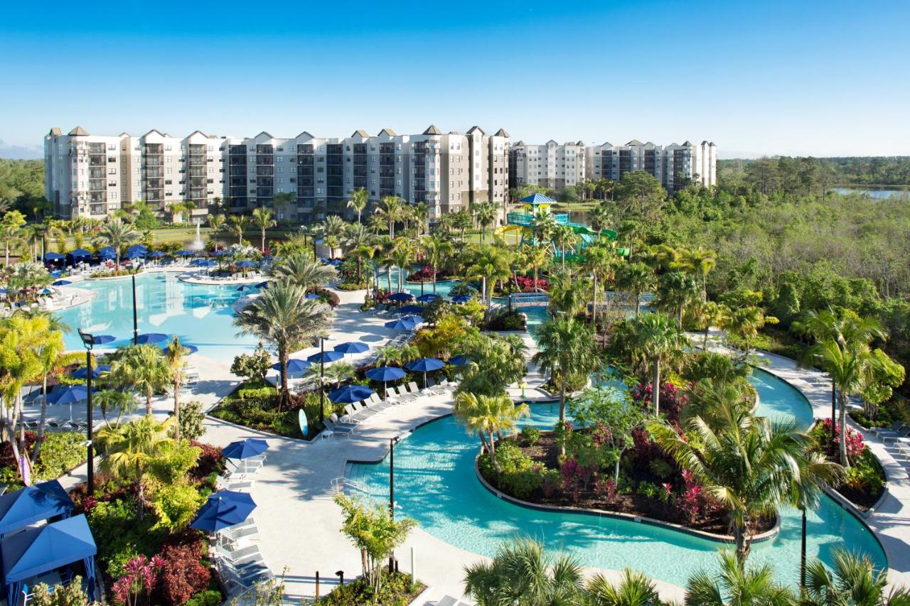 large orlando resort in the back ground with many pools and water park features in front of the hotel, there is a lot of trees and vegetation around the water features 