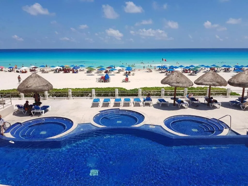 View of the luxury pool area and white sandy beach beyond. Lots of people are enjoying the beach and swimming in the turquoise waters. 
