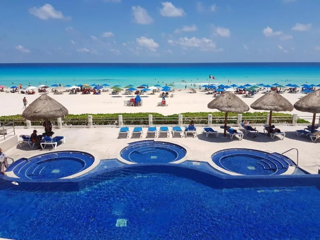 View of the luxury pool area and white sandy beach beyond. Lots of people are enjoying the beach and swimming in the turquoise waters.