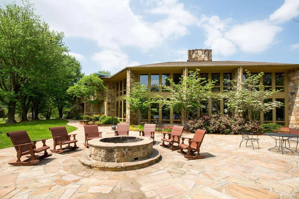 a nice outdoor seating area around a fire place and big windowed building with large trees