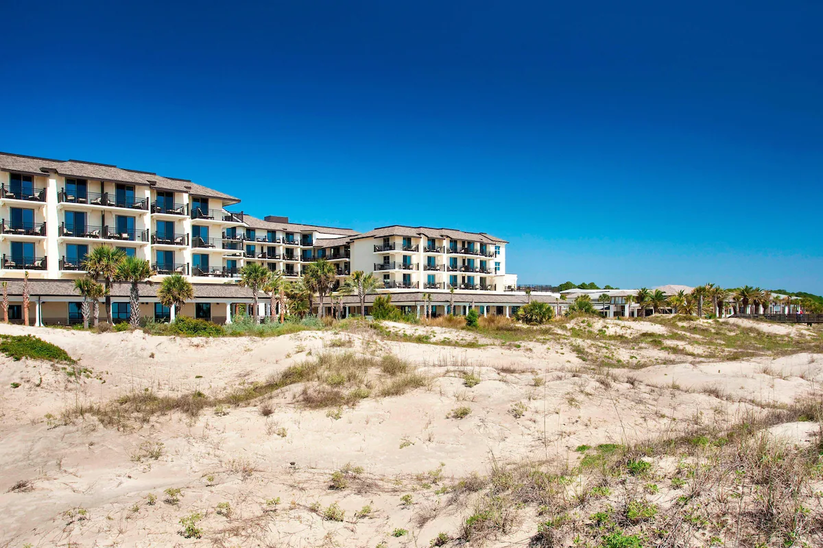 an image off the beach with grassy sprigs and sections and a lovely building complete with balconies, palm trees and with a beautifully bright blue sky behind it!